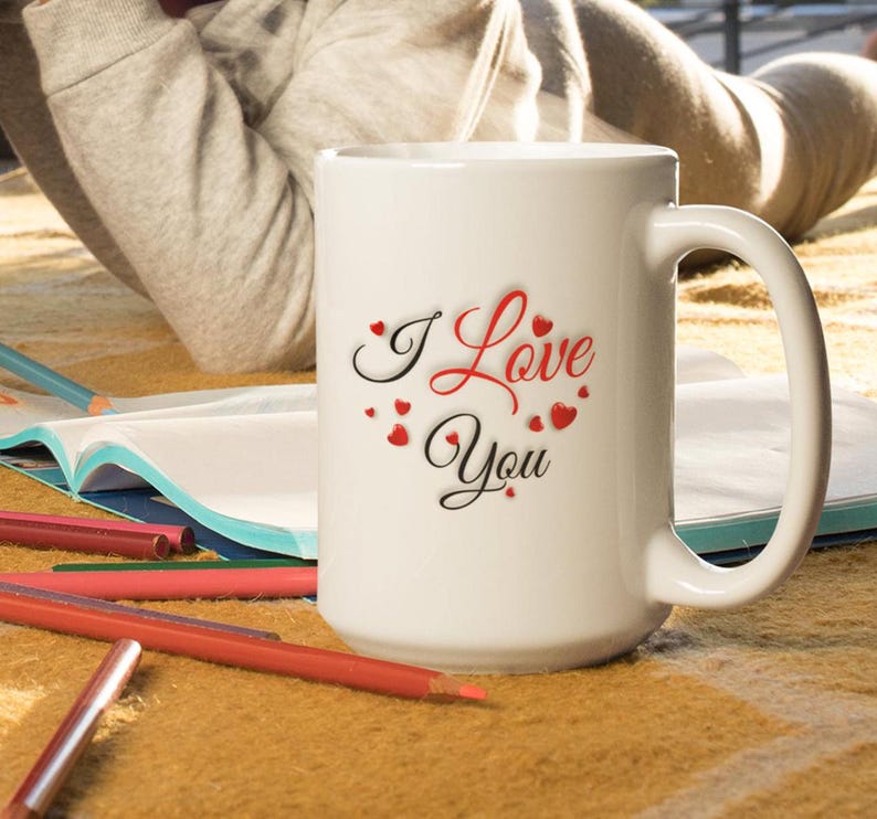 A child is lying on the floor coloring, beside her is a white ceramic mug with the words I Love You, the lettering is written in red and black letters all surrounded by many small hearts. I love you gift for him or her