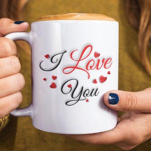 A woman is holding a white ceramic mug with the phrase I Love You in red and black lettering and is surrounded by several small, red hearts. Wonderful Valentines Day, Christmas, Birthday gift idea for him or her