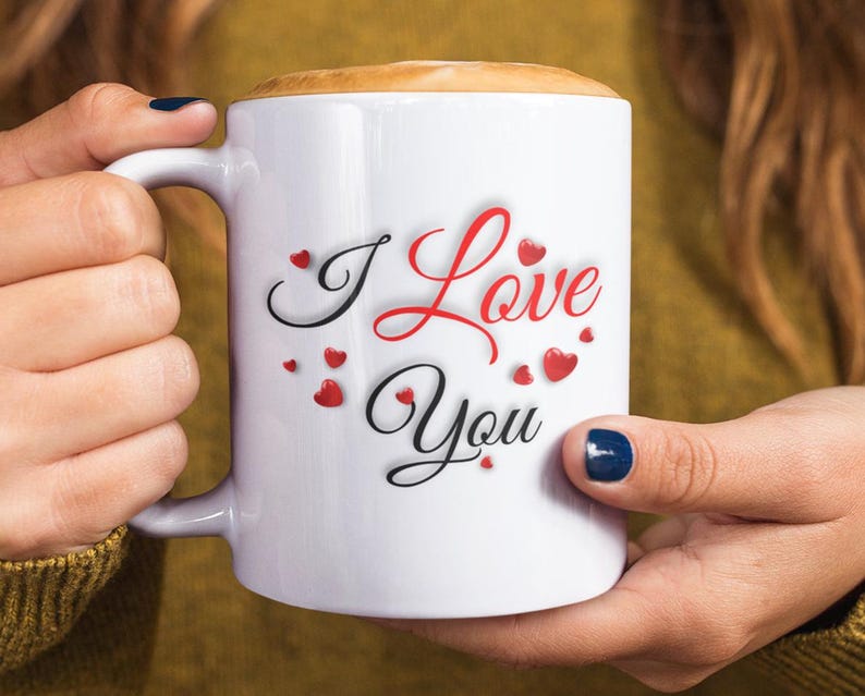 A woman is holding a white ceramic mug with the phrase I Love You in red and black lettering and is surrounded by several small, red hearts. Wonderful Valentines Day, Christmas, Birthday gift idea for him or her