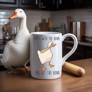 A white ceramic mug is sitting on a kitchen counter.  Next to the mug is a large white goose and a baseball bat to go along with the phrase printed on the mug, "Mess with the Honk you get the bonk".  A fun silly goose mug, great gift idea.