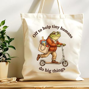 Puede incluir: Bolso tote color crema con una ilustración de una rana en un scooter. La rana lleva gafas y un suéter, llevando una bolsa con útiles escolares. El texto dice "Off to help tiny humans do big things."