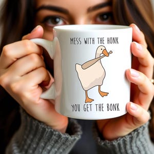 A woman is holding a white ceramic mug in both hands that is hiding most of her face.  The mug features a funny looking goose with a baseball bat in it's mouth.  The caption reads "Mess with the honk, You get the bonk".  A funny silly goose gift idea, silly gift, gag gift, dad gift.