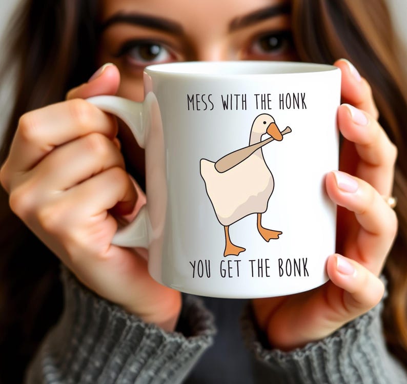 A woman is holding a white ceramic mug in both hands that is hiding most of her face.  The mug features a funny looking goose with a baseball bat in it's mouth.  The caption reads "Mess with the honk, You get the bonk".  A funny silly goose gift idea, silly gift, gag gift, dad gift.