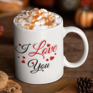 A white ceramic mug, full of hot chocolate and whipped cream, is sitting on a table with cookies and a pine cone.  The mug features the phrase I Love You with small red hearts, a wonderful Valentines Day, Christmas or Birthday gift for Mom or Dad, Boyfriend or Girlfriend.