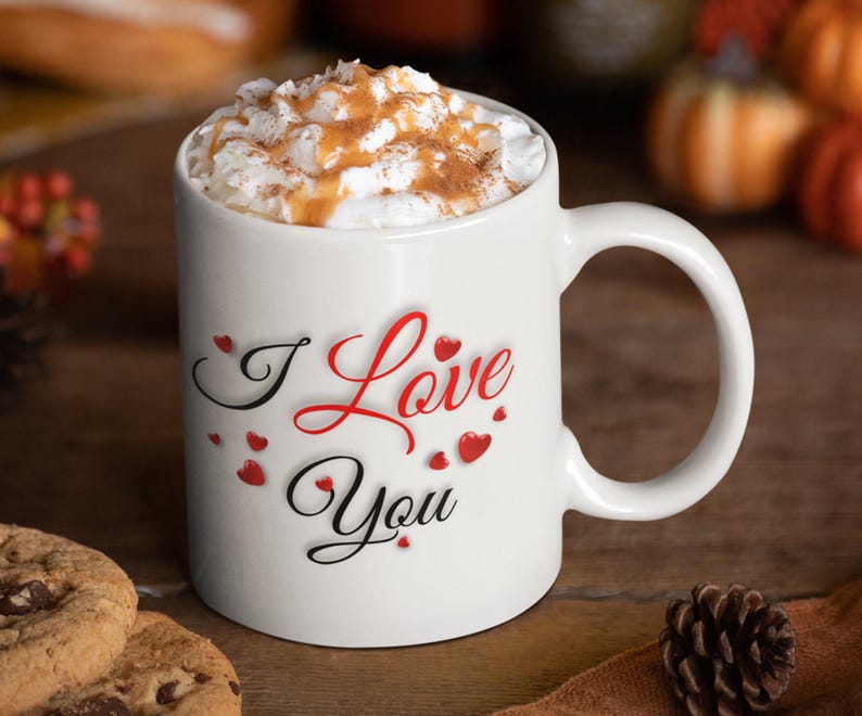A white ceramic mug, full of hot chocolate and whipped cream, is sitting on a table with cookies and a pine cone.  The mug features the phrase I Love You with small red hearts, a wonderful Valentines Day, Christmas or Birthday gift for Mom or Dad, Boyfriend or Girlfriend.