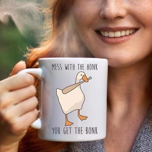 A smiling woman with red hair, is holding a white ceramic mug in her right hand.  The mug features a funny looking goose with a baseball bat in it's mouth.  The caption reads "Mess with the honk, You get the bonk".  A funny silly goose gift idea, silly gift, gag gift, dad gift.