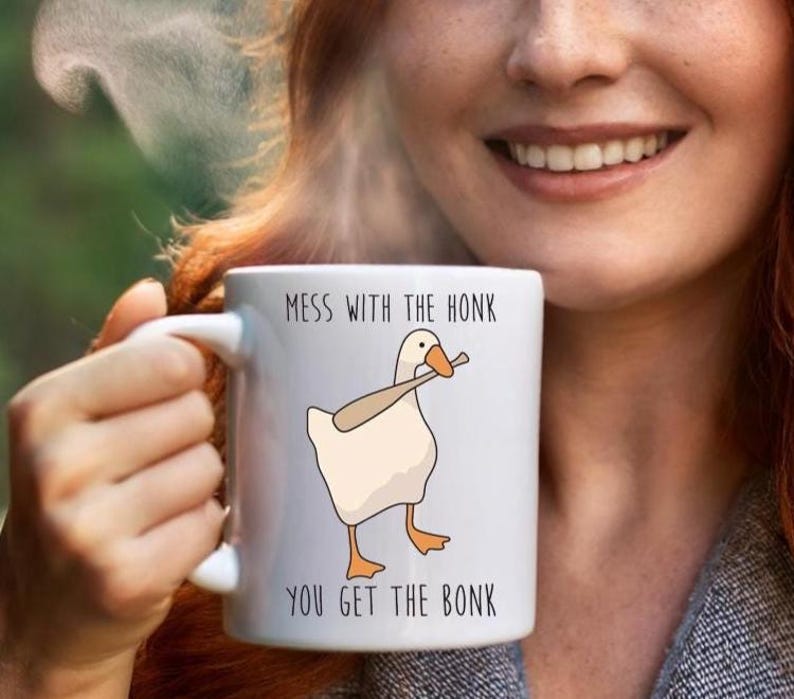 A smiling woman with red hair, is holding a white ceramic mug in her right hand.  The mug features a funny looking goose with a baseball bat in it's mouth.  The caption reads "Mess with the honk, You get the bonk".  A funny silly goose gift idea, silly gift, gag gift, dad gift.