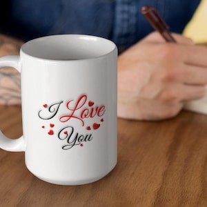 A man sitting at a table, is writing in a journal.  He has a white ceramic mug next to him.  The mug features the phrase I Love You written in red and black letters and surrounded by small hearts.  A wonderful Valentines Day, Christmas or Birthday gift for Mom or Dad, Boyfriend or Girlfriend.