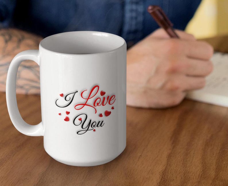 A man sitting at a table, is writing in a journal.  He has a white ceramic mug next to him.  The mug features the phrase I Love You written in red and black letters and surrounded by small hearts.  A wonderful Valentines Day, Christmas or Birthday gift for Mom or Dad, Boyfriend or Girlfriend.