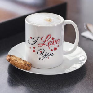 A white ceramic mug, filled with hot chocolate and whipped cream, is sitting on a small saucer with a chocolate chip cookie next to it.  The mug has I Love You written in red and black letters and surrounded by small hearts. Valentines Day Gift