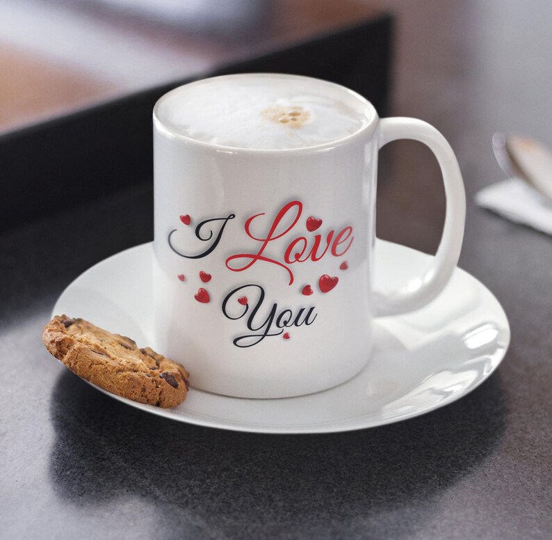 A white ceramic mug, filled with hot chocolate and whipped cream, is sitting on a small saucer with a chocolate chip cookie next to it.  The mug has I Love You written in red and black letters and surrounded by small hearts. Valentines Day Gift
