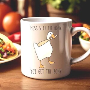 A white ceramic mug is sitting on a dining room table, behind the mug are large plates of food.  The mug features a funny looking goose with a baseball bat in it's mouth.  The caption reads "Mess with the honk, You get the bonk".  A funny silly goose gift idea, silly gift, gag gift, dad gift.