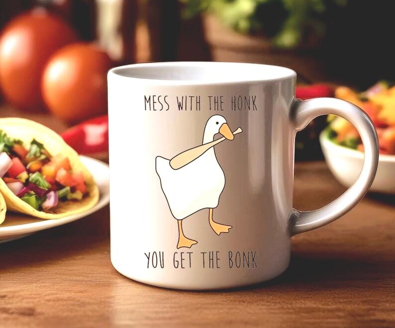 A white ceramic mug is sitting on a dining room table, behind the mug are large plates of food.  The mug features a funny looking goose with a baseball bat in it's mouth.  The caption reads "Mess with the honk, You get the bonk".  A funny silly goose gift idea, silly gift, gag gift, dad gift.