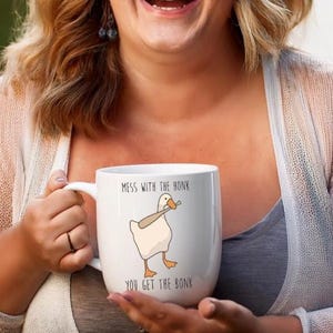 A laughing woman is holding a white ceramic mug with on hand on the handle and one hand supporting the bottom.  The mug features a funny looking goose with a baseball bat in it's mouth.  The caption reads "Mess with the honk, You get the bonk".  A funny silly goose gift idea, silly gift, gag gift, dad gift.