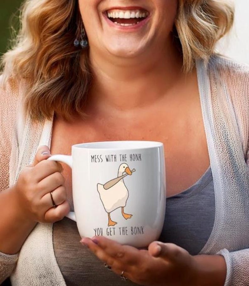 A laughing woman is holding a white ceramic mug with on hand on the handle and one hand supporting the bottom.  The mug features a funny looking goose with a baseball bat in it's mouth.  The caption reads "Mess with the honk, You get the bonk".  A funny silly goose gift idea, silly gift, gag gift, dad gift.