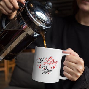 A woman is pouring coffee into a white ceramic mug that features the heartwarming phrase I Love You that is written in red and black letters all surrounded by many small hearts.  A wonderful gift to show love and appreciation, a wonderful Valentines Day, Christmas or Birthday gift for Mom or Dad, Boyfriend or Girlfriend.
