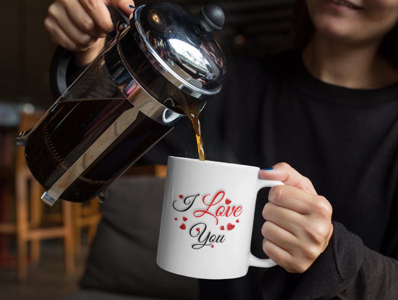 A woman is pouring coffee into a white ceramic mug that features the heartwarming phrase I Love You that is written in red and black letters all surrounded by many small hearts.  A wonderful gift to show love and appreciation, a wonderful Valentines Day, Christmas or Birthday gift for Mom or Dad, Boyfriend or Girlfriend.