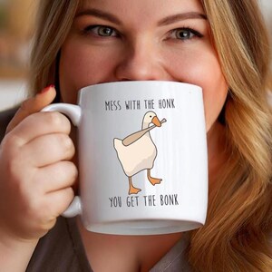 A woman is holding a white ceramic mug that is hiding most of her face.  The mug features a funny looking goose with a baseball bat in it's mouth.  The caption reads "Mess with the honk, You get the bonk".  A funny silly goose gift idea, silly gift, gag gift, dad gift.