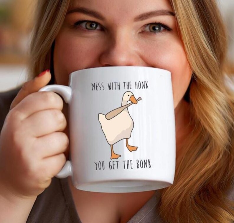 A woman is holding a white ceramic mug that is hiding most of her face.  The mug features a funny looking goose with a baseball bat in it's mouth.  The caption reads "Mess with the honk, You get the bonk".  A funny silly goose gift idea, silly gift, gag gift, dad gift.