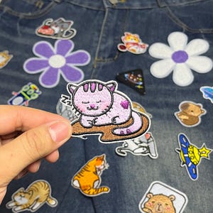 Peut inclure: Un écusson thermocollant brodé représentant un chat rose clair endormi avec des rayures violettes et une tache en forme de cœur sur le dos, reposant sur une surface brune avec la lettre Z répétée trois fois, sur un fond en tissu denim.