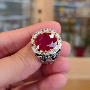 Può includere: Un anello d'argento con un grande rubino rosso circondato da pietre bianche più piccole. L'anello presenta un design floreale dettagliato.