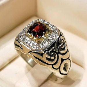 Könnte beinhalten: Ein silberner Ring mit einem roten Edelstein, der von kleineren weißen Edelsteinen umgeben ist. Der Ring hat ein detailliertes Design mit komplizierten Verzierungen.