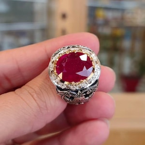 Peut inclure: Une bague ornée d'un rubis de forme ovale. La bague est sertie dans un anneau en or et argent avec des détails de filigrane complexes. De petites pierres précieuses claires entourent le rubis. La bague est tenue dans une main.