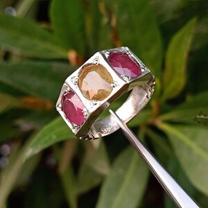 Pode incluir: Um anel de prata com três pedras preciosas ovais. A pedra central é amarela e as duas pedras de cada lado são vermelhas.
