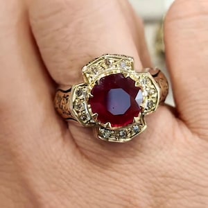 Puede incluir: Un anillo de oro con una gran piedra de rubí roja rodeada de diamantes más pequeños. El anillo tiene detalles intrincados y un diseño en forma de cruz.