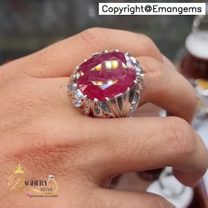 May include: A large, oval-cut ruby gemstone ring. The ruby is a deep red color and is set in a silver band with ornate detailing. The ring is a statement piece, perfect for special occasions.