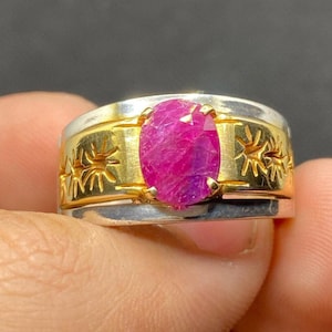 May include: A silver and gold ring featuring a pink oval ruby gemstone. The ring band has a decorative design.