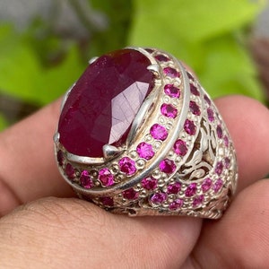 Może przedstawiać: Srebrny pierścionek z dużym, owalnym rubinem otoczonym mniejszymi rubinami. Pierścionek ma skomplikowany wzór z motywami kwiatowymi.