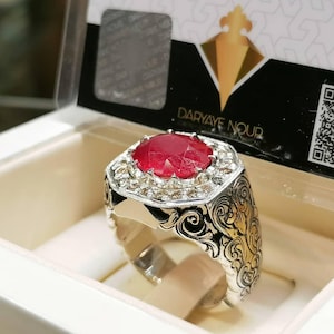 Könnte beinhalten: Ein silberner Ring mit einem großen, roten Rubin-Edelstein, der von kleineren, weißen Edelsteinen umgeben ist. Der Ring hat ein filigranes, florales Design auf der Ringschiene. Der Ring befindet sich in einer weißen Schachtel mit dem Text "DARYAYE NOOR" auf dem Deckel.