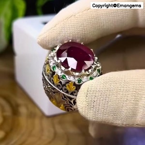 May include: A silver ring featuring a large, oval-cut ruby gemstone. The ring is adorned with smaller white and green gemstones, and gold and black filigree detailing. The ring is held by a gloved hand.