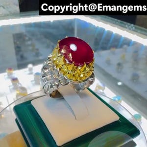 May include: An elaborate ring with a striking red gemstone. The ring has a gold-coloured setting around the stone and a silver-coloured band with detailed patterns. The text "Copyright@Emangems" is visible at the top.