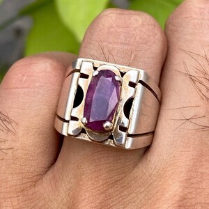 Op de afbeelding: Een zilveren ring met een grote, ovale, paarse edelsteen die is gezet in een gedetailleerd, geometrisch ontwerp.
