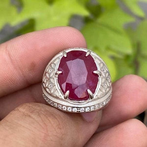 May include: A silver ring with a large, oval-cut ruby gemstone. The ruby is a deep red hue, set in a detailed silver band with small, clear accent stones. The ring is displayed against a blurred green backdrop.