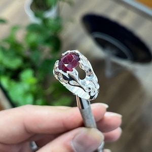 May include: A silver ring with an oval, deep red gemstone. The ring features an ornate, flowing design around the gem. The ring is held by a tool, with a blurred background of greenery.
