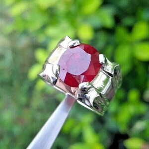 May include: A silver ring featuring a large, round, red gemstone set in the centre. The ring has a detailed, textured band.