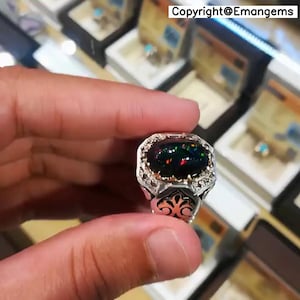 May include: A silver ring featuring an oval black opal gemstone. The opal displays flashes of green, red, and blue. The ring has a detailed filigree design around the stone and on the band. The text "Copyright@Emangems" is visible in the background.