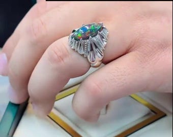 May include: A silver ring featuring an oval, iridescent black opal gemstone. The ring has an elaborate, ornate setting with a textured design. The text "Copyright@EmanGems" is visible in the image.
