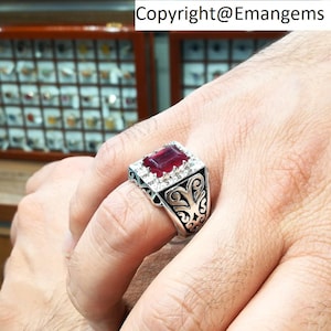 May include: A silver ring featuring a large, square, red gemstone surrounded by smaller white gemstones. The ring has an intricate, ornate design.