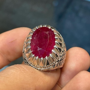 Peut inclure: Une bague en argent avec une grande pierre précieuse de rubis rouge de forme ovale sertie dans un décor orné et détaillé.
