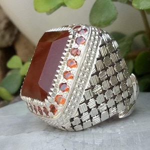 Peut inclure: Une bague en argent avec une grande pierre rectangulaire brun rougeâtre sertie au centre. La bague est décorée d'une bordure de petites pierres rouges et d'une bande texturée et à motifs.