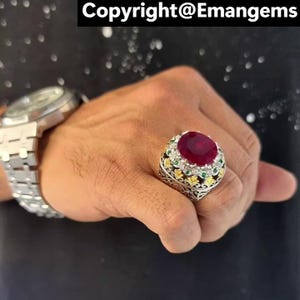 Peut inclure: Une bague en argent ornée d'un grand rubis ovale. La bague est ornée de petites pierres vertes et jaunes. Le texte "Copyright@Emangems" est visible en haut.