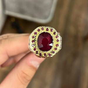 Puede incluir: Un anillo de oro con una gran piedra preciosa roja rodeada de piedras preciosas rojas más pequeñas.