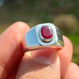 Puede incluir: Un anillo de plata con una piedra preciosa roja engastada en un engaste de bisel. El anillo tiene una banda ancha y un acabado liso.