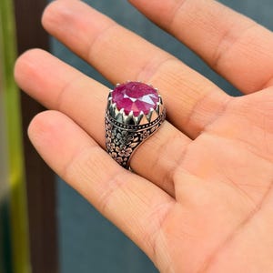 May include: A silver ring featuring a large, faceted ruby gemstone. The ring has a floral pattern on the band and a crown-shaped setting around the ruby. The image includes the text "Copyright@Emangems".