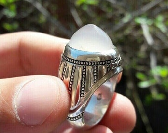 May include: A silver ring with a white gemstone set in a decorative bezel. The ring has a textured band with a repeating pattern of small circles.