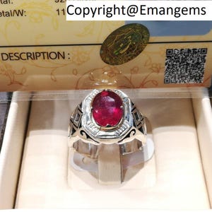 Pode incluir: Um anel de prata com uma grande pedra de rubi de corte oval. O anel tem um design geométrico detalhado na banda e uma moldura de metal branco ao redor do rubi. O texto "Copyright@Emangems" é visível.